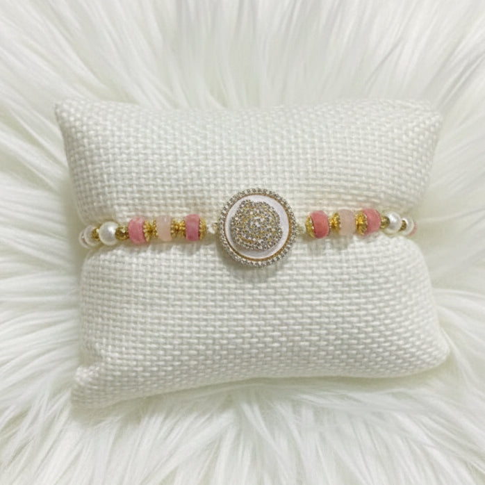 A beaded bracelet displayed on a textured white pillow. The bracelet features pink and white beads, gold spacer beads, and a central round silver-toned medallion embellished with small clear stones.